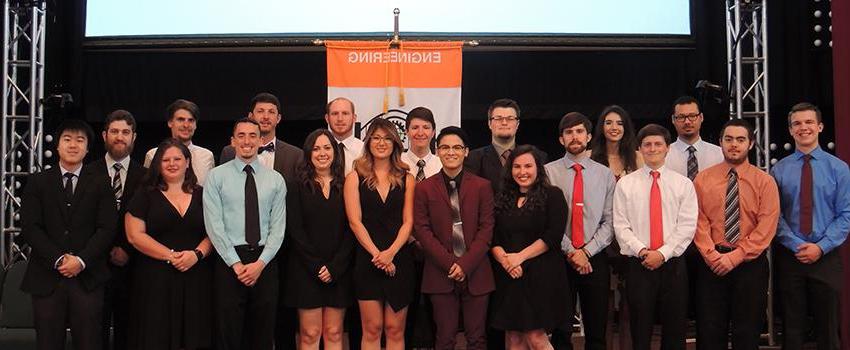 Engineering students standing on stage in front of flag.